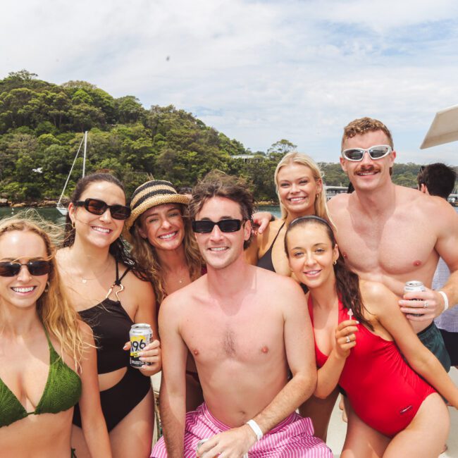 A group of seven young adults smiling and posing on a boat, wearing swimsuits and sunglasses, with tropical trees and water in the background. Some hold drinks, and the atmosphere is sunny and cheerful.