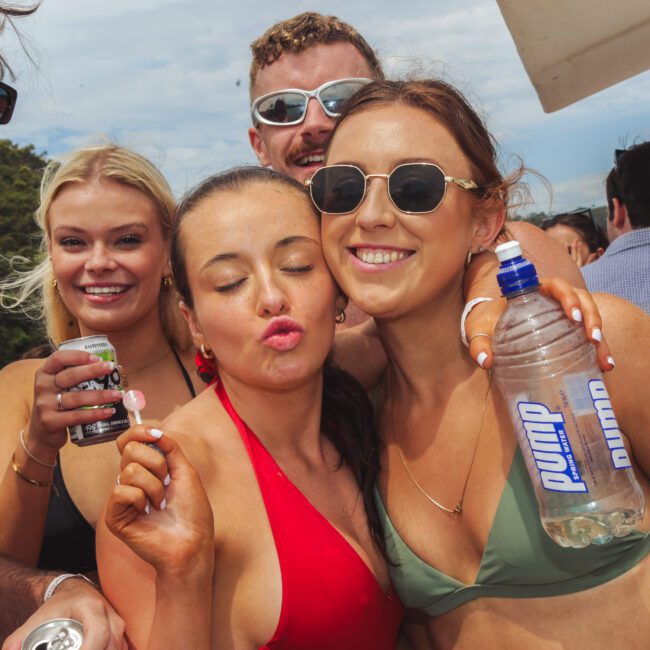 A group of young adults in swimsuits smile and pose closely together at an outdoor party; one woman holds a water bottle, another holds a drink, and they all appear to be enjoying a sunny day.