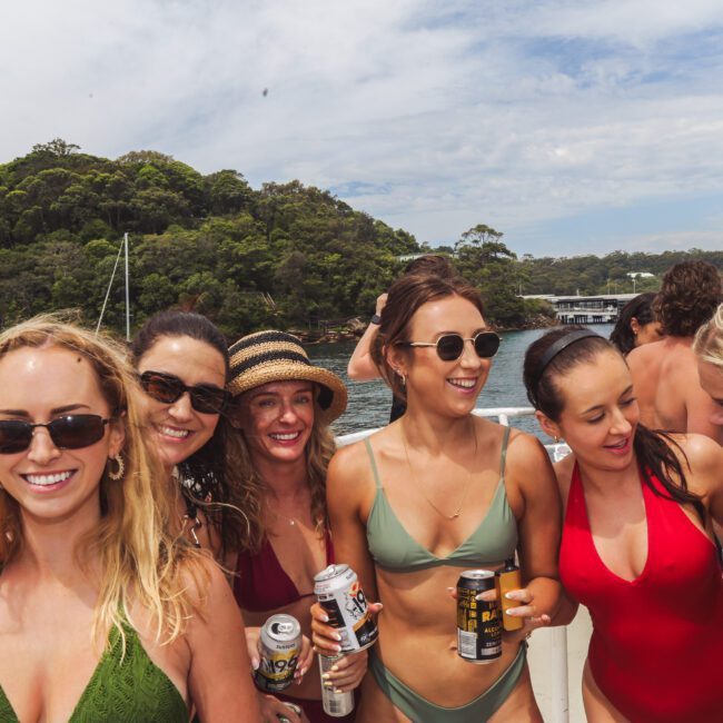 A group of six women in swimsuits smile and pose together on a boat, holding drinks, with trees, water, and sailboats in the background on a sunny day.