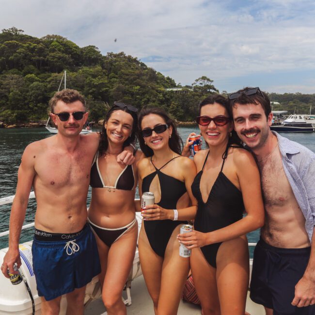Five young adults in swimwear stand close together, smiling and holding drinks on a boat, with forested hills, anchored boats, and blue water visible in the background.
