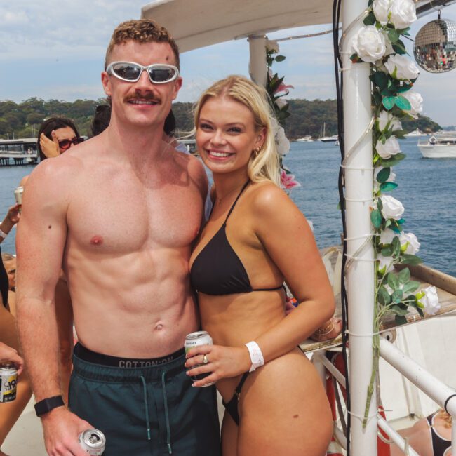 A smiling man and woman in swimsuits pose together on a decorated boat with drinks in hand. Other people are mingling in the background, and boats are visible on the water under a sunny sky.