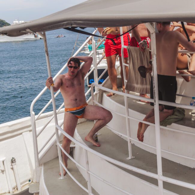A man in orange swim briefs poses playfully on the steps of a boat, holding onto the railing, while a group of people in swimsuits relax and chat on the upper deck with the ocean and other boats in the background.