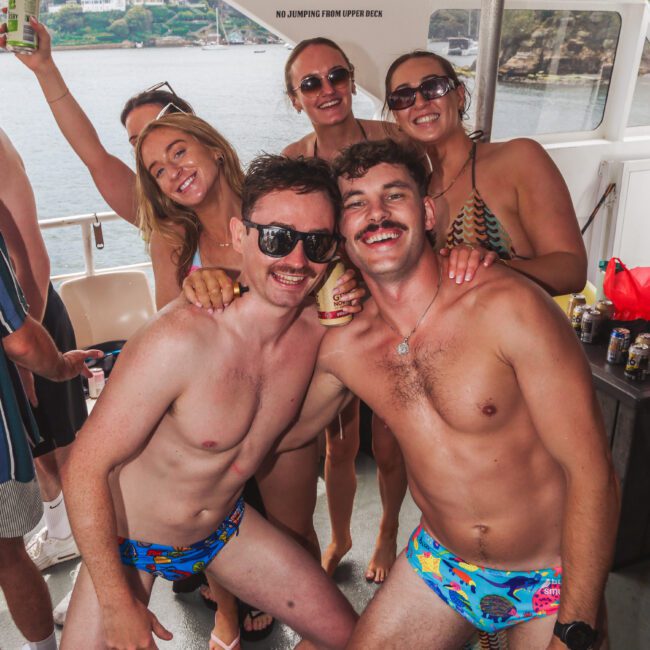 A group of smiling people in swimsuits pose together on a boat, with two men in colorful swim briefs kneeling at the front. The background shows water and land. Everyone looks happy and cheerful.