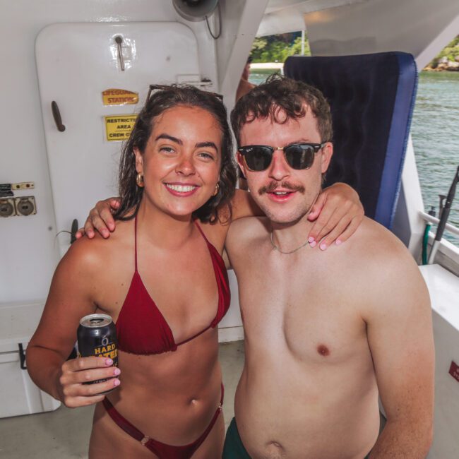 Two smiling people in swimsuits pose together on a boat. The woman holds a can and the man wears sunglasses. Water and trees can be seen in the background through the boat's railing.