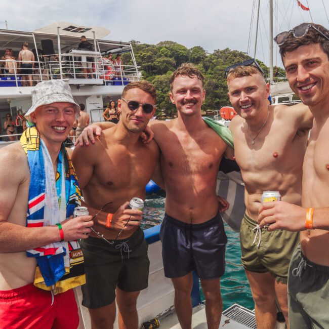 Five smiling men in swim trunks stand arm-in-arm on a boat, holding drinks. Other boats and people are visible in the background, with green trees and blue water surrounding them. It’s a sunny day.