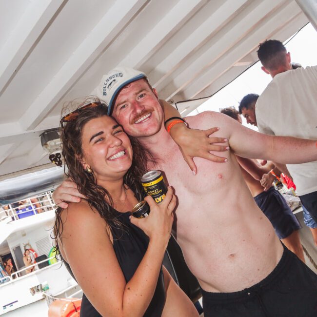 A smiling woman in a black swimsuit and a smiling shirtless man with a mustache pose together on a boat. The woman holds a drink can, and other people are visible in the background, enjoying the boat party.