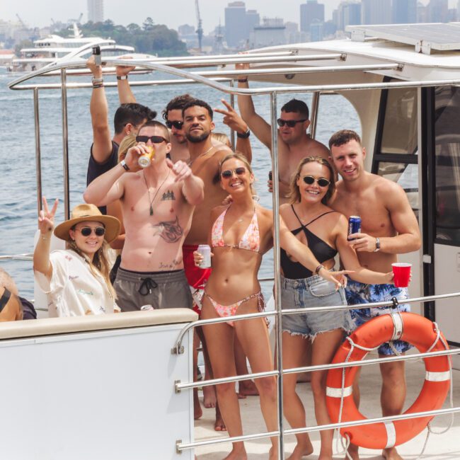 A group of young adults in swimsuits and sunglasses enjoy drinks and pose for a photo on a boat, with water and a city skyline in the background. Some smile and make peace signs, and the weather appears sunny.