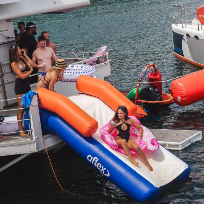 A group of people on a boat enjoy the water; a woman in a black swimsuit slides down an inflatable slide into the sea while others watch and relax nearby with colorful floaties.
