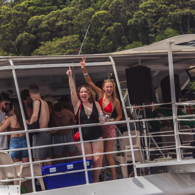 A group of people enjoy a lively party on a boat; two women in front, smiling and cheering, hold their arms up while others mingle and relax in swimwear. Lush green trees are visible in the background.