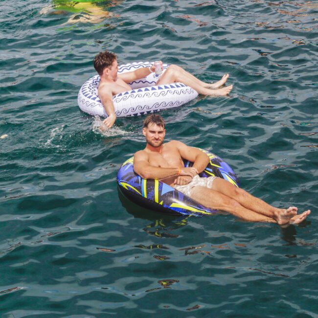 Two men float on inflatable tubes in a large body of water. The man in front is on a dark patterned tube and smiling at the camera, while the man behind him relaxes on a white tube with a blue design.