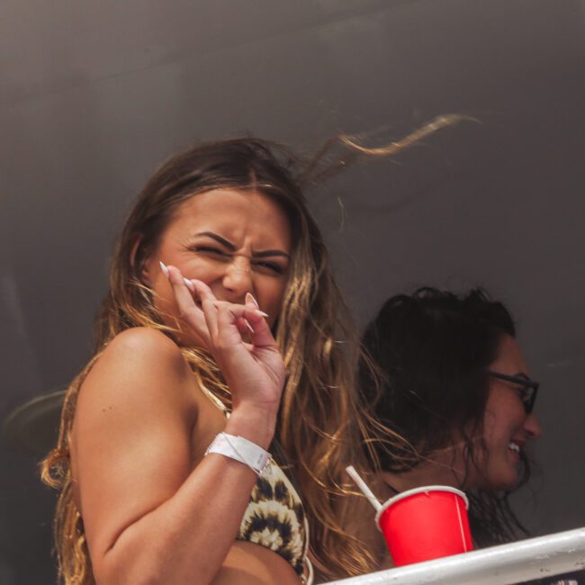 A woman in a swimsuit stands on a balcony, squinting and making a playful face while holding a red cup. Another person with long dark hair smiles in the background. The scene looks casual and festive.