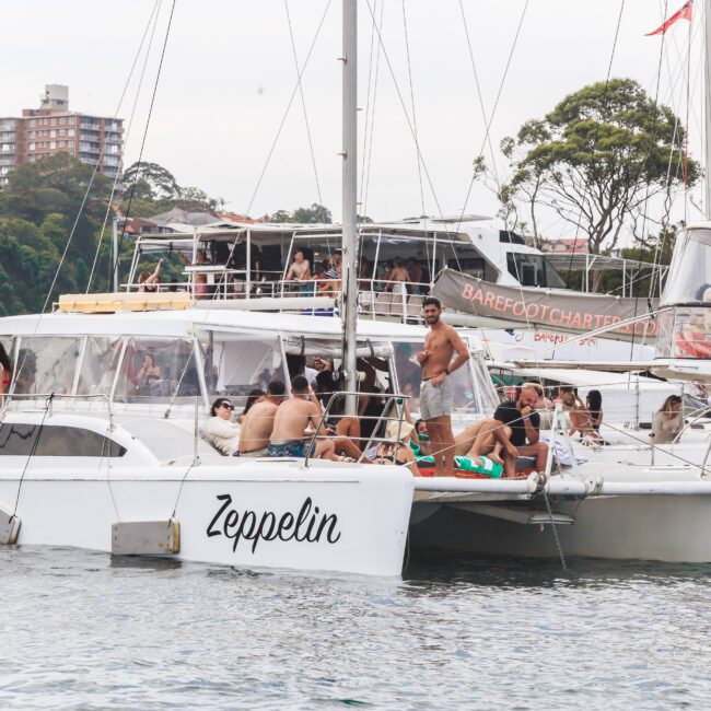 Several people relax and socialize on a white catamaran named "Zeppelin," docked on the water near other boats. Trees and a tall apartment building are visible in the background.