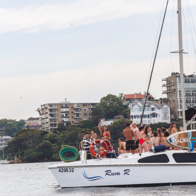 A group of people in swimwear stand and sit on the deck of a white catamaran named "Rum R," enjoying a sunny day on the water with apartment buildings and trees in the background.