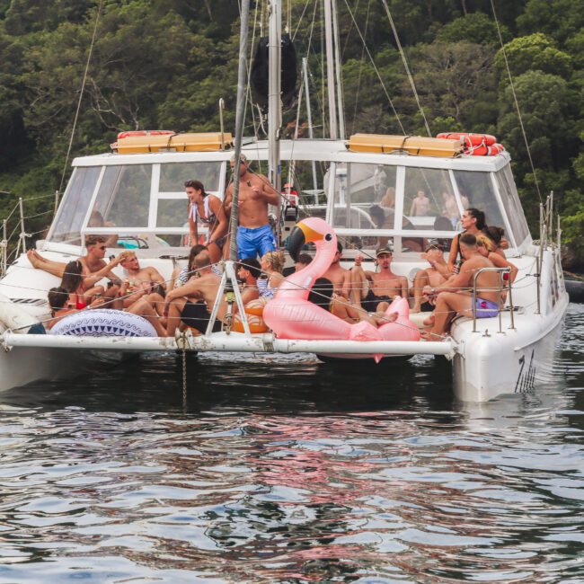 A group of people in swimsuits relax and socialize on the deck of a catamaran, with inflatable pool floats, including a large pink flamingo, surrounded by water and lush green trees in the background.