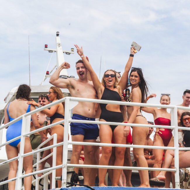 A group of young adults in swimsuits smile and cheer on the deck of a boat, enjoying a sunny day. Some wave at the camera, while others chat. The sky is partly cloudy in the background.