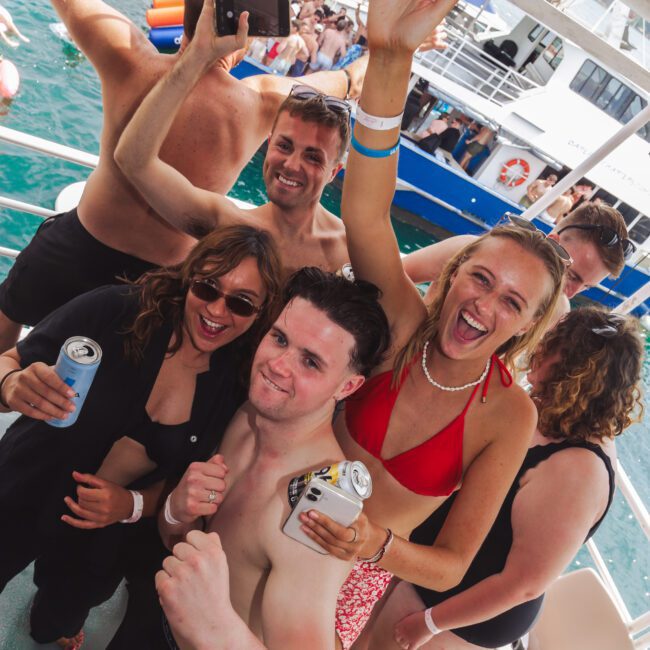 A group of people in swimsuits smile, pose with drinks, and raise their arms while on a crowded boat party under the sun, with other boats and swimmers visible on the water in the background.