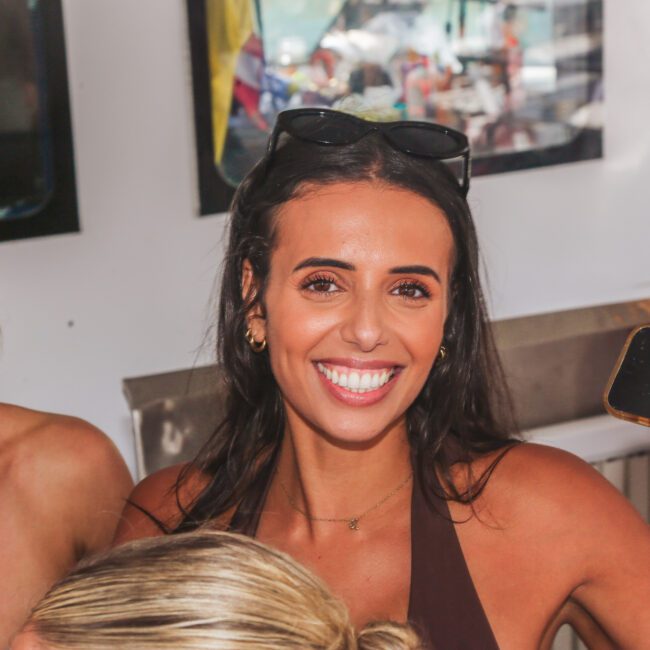 A woman with dark hair smiles brightly at the camera at a social event, holding a phone in one hand. Another woman beside her, wearing sunglasses, sips from a can. The background shows a white wall and reflections in windows.