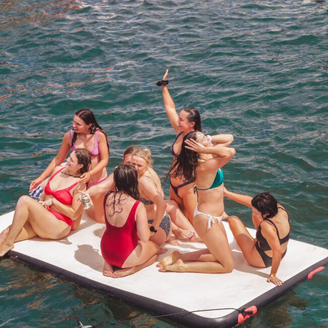 A group of women in swimsuits relax and pose on a floating mat in the water, enjoying a sunny day. One person in an orange float is nearby, with waves and splashes surrounding them.
