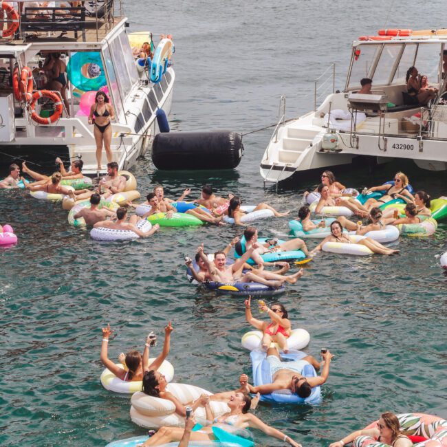Dozens of people relax on colorful inflatable floats in the water between two docked boats on a sunny day, with more people on board the boats and sailboats visible in the background.