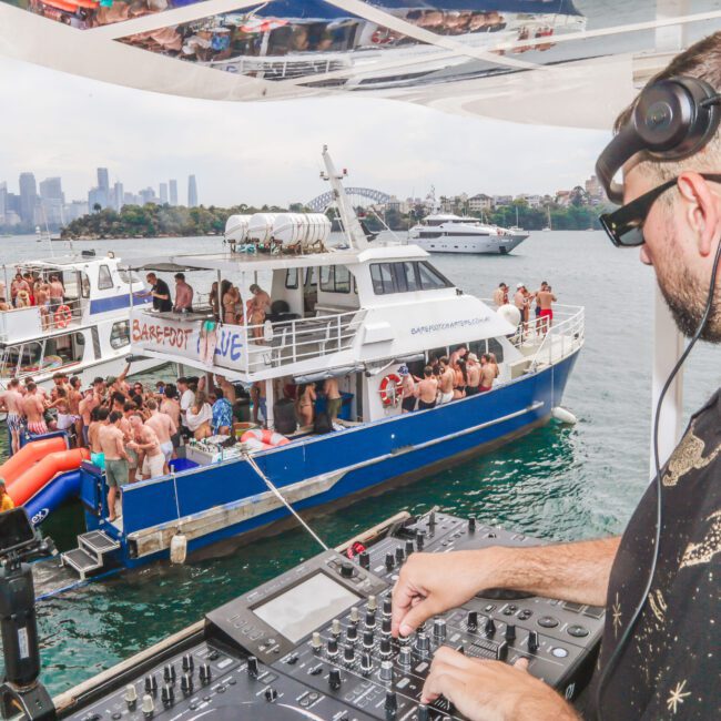 A DJ wearing sunglasses and headphones plays music on a boat deck while two crowded party boats float nearby on the water, with people dancing and city buildings visible in the background.