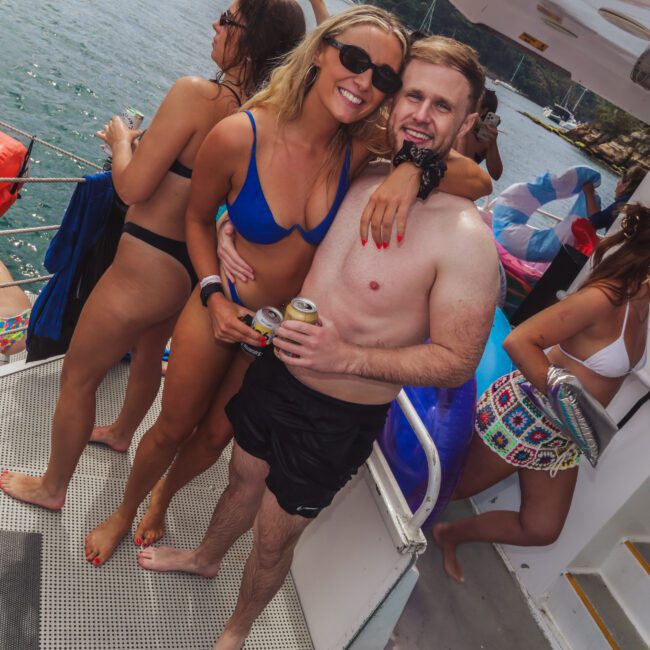 A group of people in swimwear enjoy drinks and laugh together on a boat. Two people, smiling and holding drinks, pose for a photo while others relax nearby and the water is visible in the background.