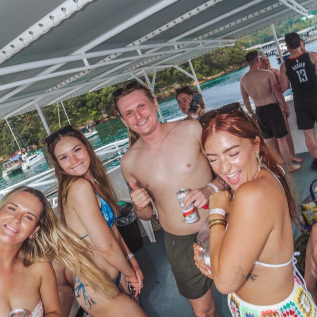 A group of young adults in swimsuits smile and pose on a boat, holding drinks. The boat is on the water with greenery visible in the background. The atmosphere appears lively and joyful.