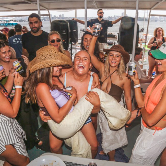 A group of people in swimwear laugh and celebrate on a boat. Several women hold drinks and wear hats; one woman lifts a smiling man in swim trunks. The boat is crowded, with water and other boats in the background.