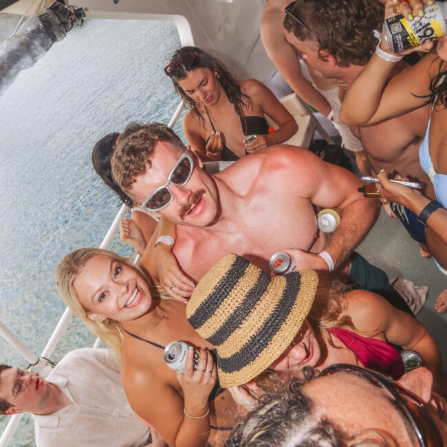A group of young adults in swimsuits and summer attire enjoy drinks and dance closely together on a boat, with sunlight streaming in and water visible outside the windows.