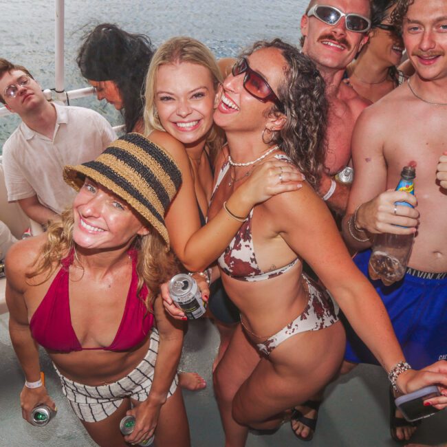 A group of young adults in swimsuits smile and pose for a photo on a boat, holding drinks, with water visible in the background. They appear to be enjoying a lively party under bright daylight.