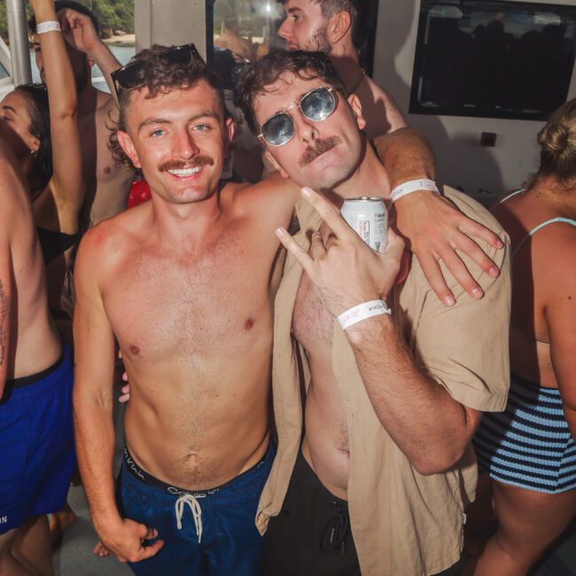 Two men with mustaches, wearing swimsuits and wristbands, pose and smile on a crowded boat party. One holds a can and makes a hand gesture, while others dance and mingle in the background.