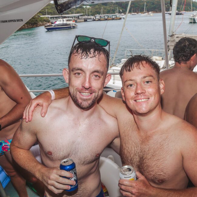 Two smiling men in swim trunks with drinks pose together on a boat, surrounded by other people in swimwear. The background shows water, yachts, and a forested shoreline under an overcast sky.