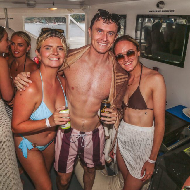 A group of young adults in swimwear smile and pose together on a boat, holding drinks. The atmosphere is lively, with several people socializing in the background. Sunlight streams through the windows.