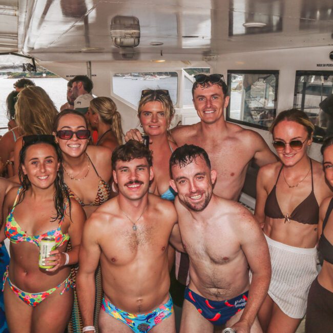 A group of young adults in swimsuits smile and pose together on a boat during the day, with water and more people visible in the background. Some are holding drinks, and the atmosphere appears lively and fun.
