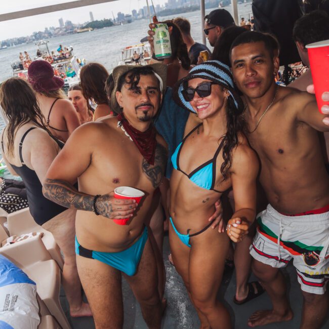 Three people in swimsuits pose and smile for a photo on a crowded party boat, with others in the background, some holding drinks. The city skyline and water are visible behind them.