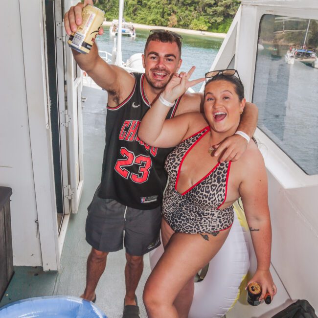 A man in a Chicago Bulls jersey and a woman in a leopard print swimsuit smile and pose with drinks on a boat, with water and greenery visible outside. An inflatable blue pool float is on the floor beside them.