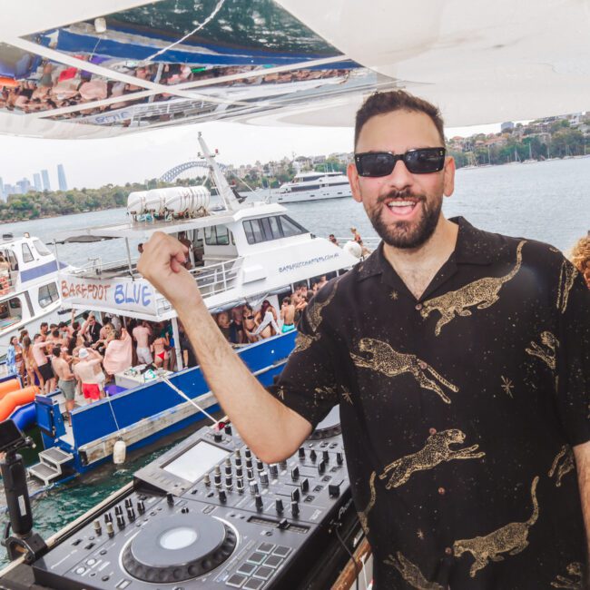 A smiling DJ in sunglasses stands by a mixing console on a boat, gesturing towards a nearby party boat full of people on the water, with a city skyline and more boats visible in the background.