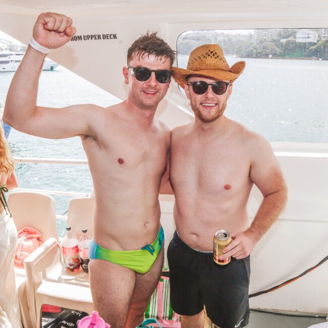 Two men in swimwear and sunglasses pose on a boat; one raises his arm, the other wears a cowboy hat and holds a drink. There are other people in swimwear and water visible through the window behind them.