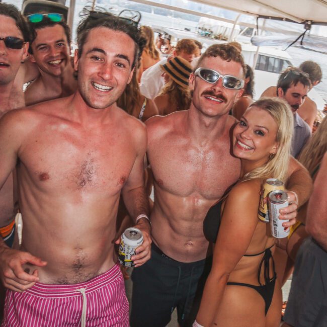 A group of young adults in swimwear smile and pose together on a boat, holding drinks. The scene is lively and sunny, with other people and water visible in the background.
