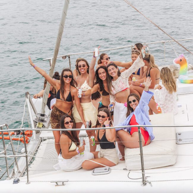 A group of women in swimwear and summer outfits smile, pose, and celebrate on a boat deck on the water, holding drinks with a pool float and the ocean visible in the background.