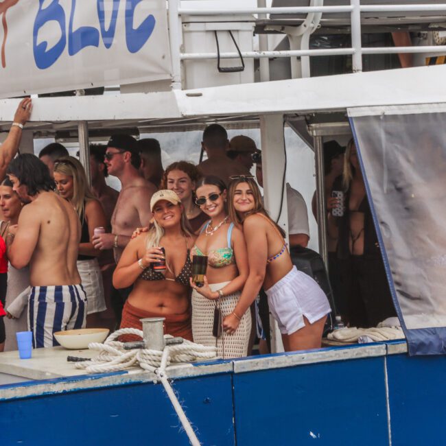 A group of young people in swimsuits are socializing and smiling on the deck of a boat. Three women pose for a photo in the foreground, while others mingle and relax behind them. The boat has a sign reading "Barefoot Blue.