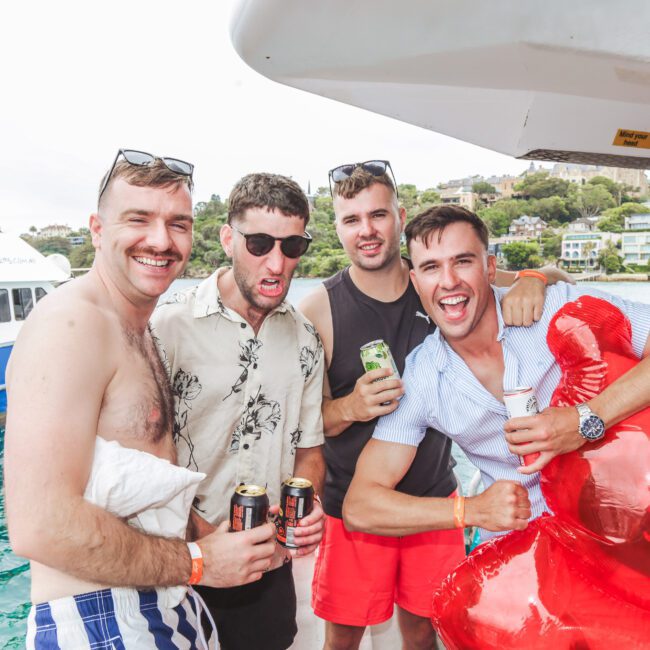 Four young men smile and pose together on a boat, holding drinks. The background shows water, a white ferry, and houses on a hill. One man holds an inflatable red float. The mood is fun and relaxed.