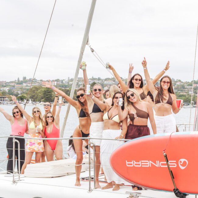 A group of women in summer outfits stand and pose cheerfully on a sailboat, smiling and raising their arms. The boat is docked near a shoreline with houses and trees visible in the background.
