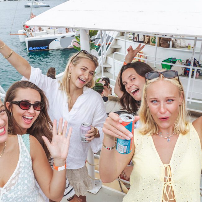 Five young women smiling, laughing, and holding drinks, pose together on a boat under a partly cloudy sky. Other people and boats are in the background, creating a lively and festive atmosphere.