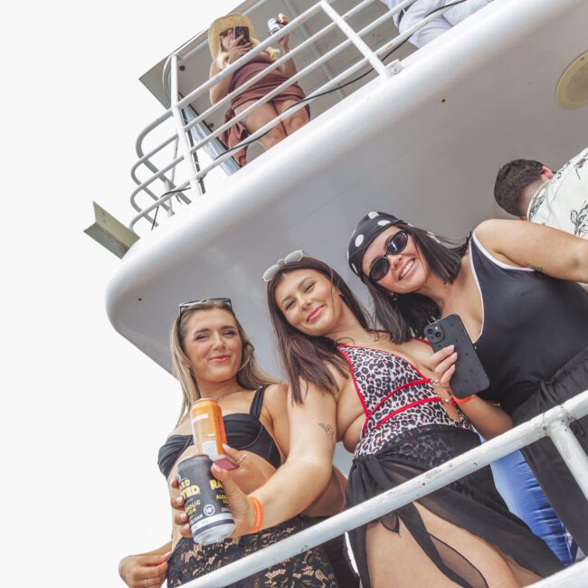 Three women smiling and posing for a photo on a boat, holding drinks. Other people are seen in the background on the upper and lower decks. The atmosphere appears relaxed and festive.