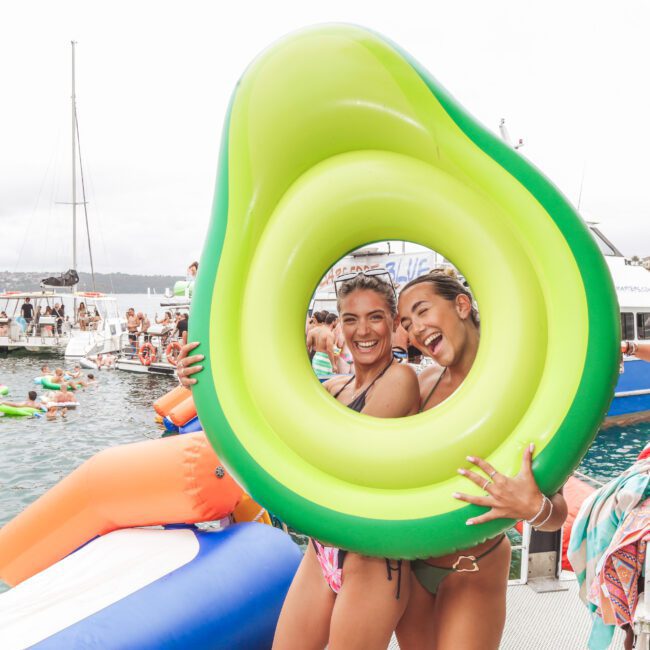 Two women in swimsuits smile and pose together through a giant inflatable avocado at a lively dockside pool party, with people, boats, and more floaties in the background.