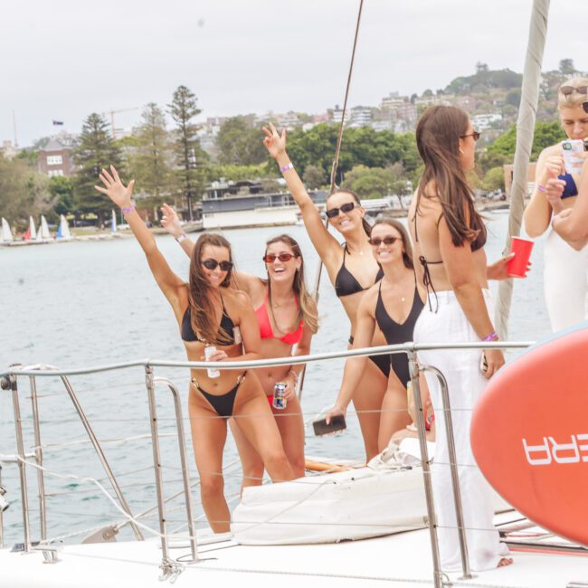 A group of women in swimsuits smile and wave on a yacht, enjoying a sunny day on the water. Some hold drinks, and others pose for the camera. Land with trees and buildings is visible in the background.