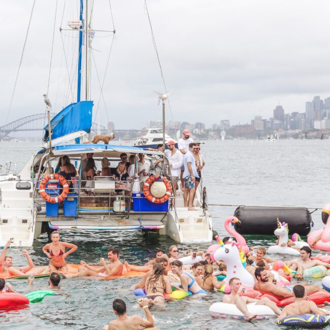 A lively crowd of people on colorful pool floats and inflatables enjoy a party in the water near boats, with a city skyline and bridge visible in the background under a cloudy sky.