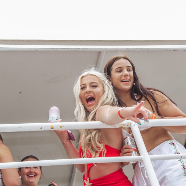 Two young women in swimwear stand on a balcony, smiling and laughing. One points excitedly while the other holds a drink can. Two more people are partially visible in the background. The atmosphere appears joyful and fun.