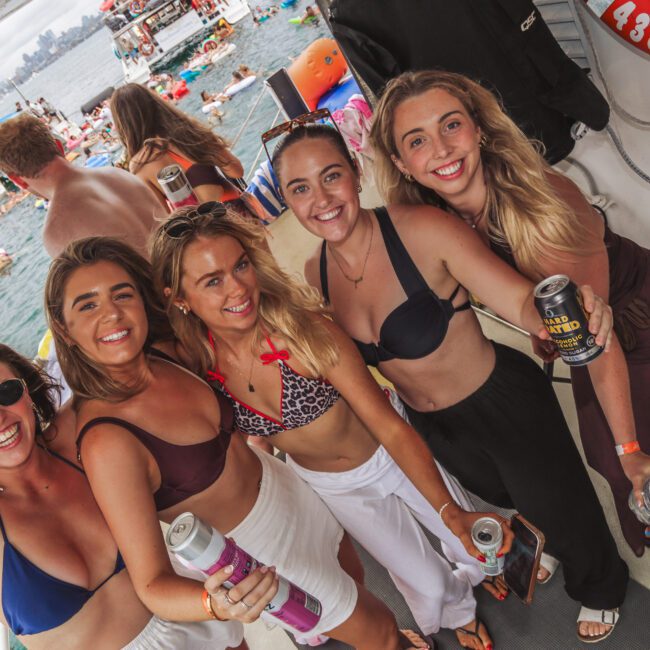 Five smiling women in swimwear pose together on a boat, holding drinks. The background shows a busy scene with other people, boats, and inflatables on the water, suggesting a lively party atmosphere.