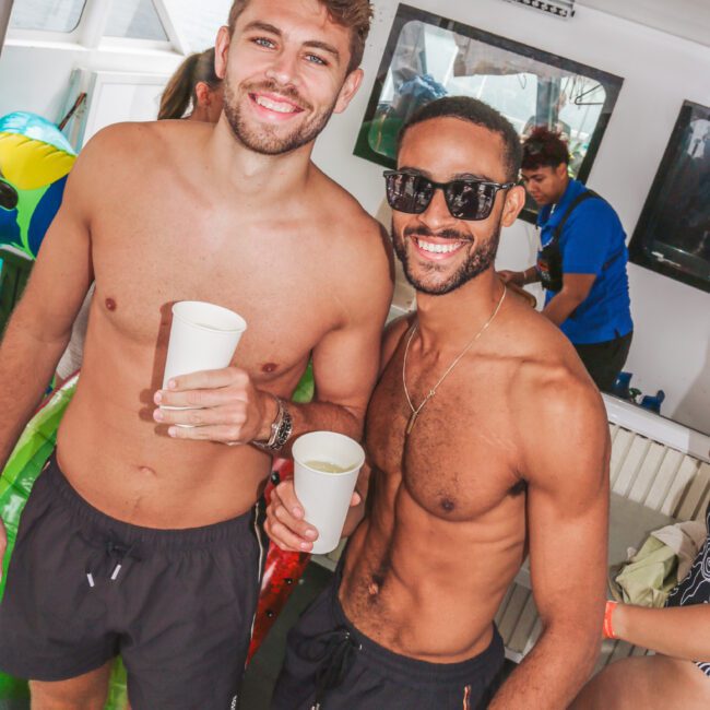 Two men in swim trunks and sunglasses smile while holding drinks on a boat. Other people and pool inflatables are visible in the background, suggesting a casual, sunny boat party atmosphere.